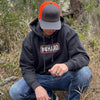 Man pictured from front wearing a WW HARD Charcoal Grey Hoodie. WW HARD "patch-style" icon printed in neon orange and white, smaller on center chest. Banded cuffs and waist. (Also wearing a WW HARD Grey/Neon Orange Retro Trucker hat.)