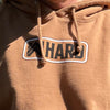 Premium hooded sweatshirt in Saddle Brown color—Front Cropped-in view of the center chest imprint. WW HARD icon with "rope and stitch" border printed on center chest in white and black ink ("patch" style.) Thick drawstring in hood, metal eyelets.