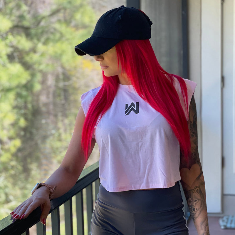 Female model facing forward but head turned away (black ball cap on), from waist up, with right arm resting on deck rail. She's wearing a women’s cropped muscle tee in pale pink. Screen-printed graphic on back includes a female "skull-style" painted face with long hair, a rose in hair over ear and tattooed “web” on shoulder. Circular text "NO EXCUSES • NEVER NOT WORKIN" printed around graphic in black ink. All text in Medieval-style BOLD font.