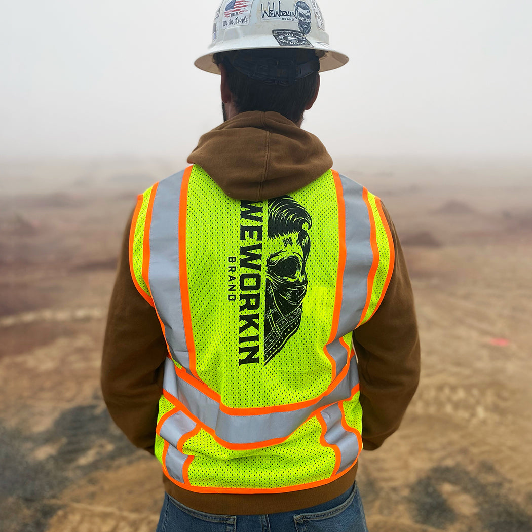 Man pictured from the back on a construction site with a hard hat on, wearing the We Workin Class 2 Mesh Zippered Two-Tone Vest. Back is printed vertically with the WEWORKIN BRAND Half skull with bandana in black ink. (Front is blank)