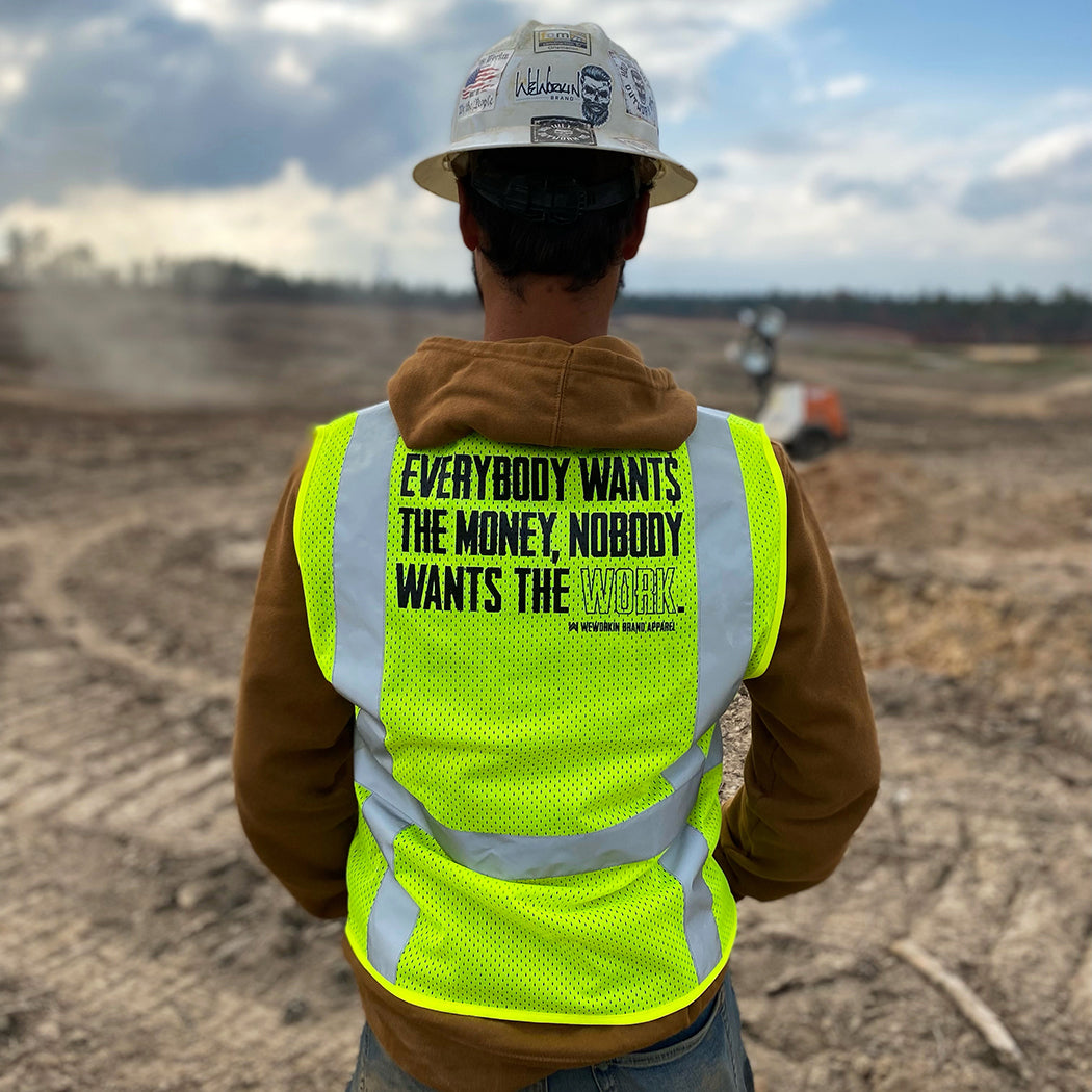 Man on a construction site, pictured from the back with a hard hat on, wearing the We Workin Class 2 Mesh Zippered 6-Pocket Vest. Back is printed with the WEWORKIN BRAND "Everybody Want$ the Money, Nobody Wants the WORK." slogan in black ink. (Front is blank)