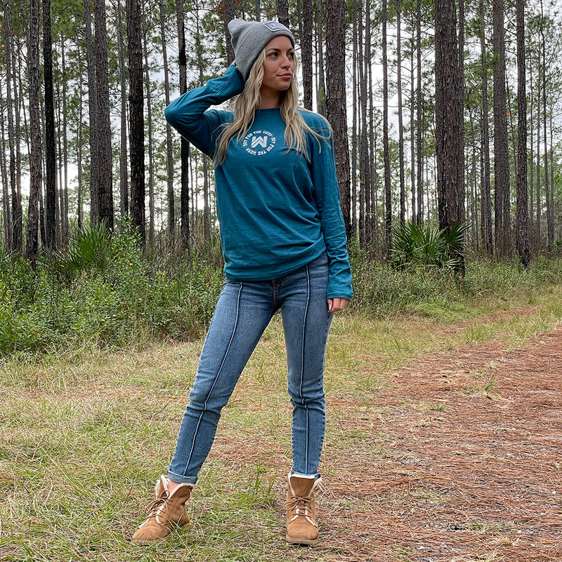 Woman pictured from front in heather teal long-sleeved tee, imprinted with the We Workin icon centered on the chest with the text "Not For The Weak" encircling the icon in white ink. Also wearing a We Workin heather grey patch beanie.