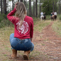 Woman kneeling, pictured from back in heather red long-sleeved tee, imprinted with the text "The Dream Never Leaves" and a dreamcatcher/indian graphic in white ink on full back.