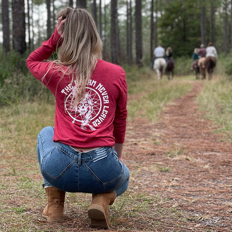 Woman kneeling, pictured from back in heather red long-sleeved tee, imprinted with the text "The Dream Never Leaves" and a dreamcatcher/indian graphic in white ink on full back.