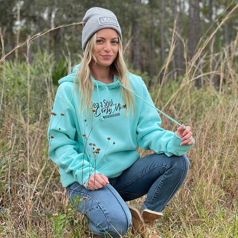 Woman pictured from front, kneeling in woods. She is wearing the medium-weight mint teal long-sleeve hooded sweatshirt. Printed with the tagline "Clean Soul. Dirty Mind.", centered on the front upper chest in black ink. (Also wearing a WEWORKIN Brand Grey Beanie)