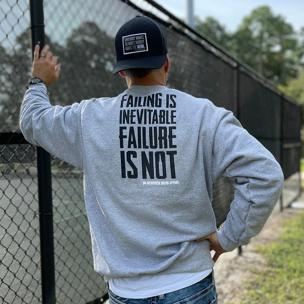 Man outdoors pictured from back wearing a WW Athletic Grey crewneck sweatshirt. "FAILING IS INEVITABLE—FAILURE IS NOT" printed high on back in black ink [WEWORKIN BRAND APPAREL printed small and lower right just below the word NOT.) Banded cuffs and waist. (Also wearing a backwards WW Retro Trucker hat with large merrowed edge patch "Everybody Wants the Money, Nobody Wants the WORK." embroidered in grey and white thread on patch.)