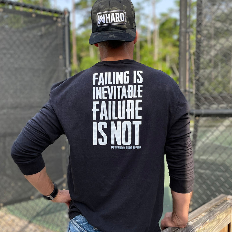 Man outdoors pictured from back wearing a WW black long sleeve shirt. "FAILING IS INEVITABLE—FAILURE IS NOT" printed high on back in white ink [WEWORKIN BRAND APPAREL printed small and lower right just below the word NOT.) Banded cuffs. (Also wearing a backwards WW Black Camo FlexFit hat with the WW HARD patch-style graphic embroidered in white and grey thread across the (2) center front panels.