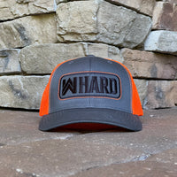 Front view of a Charcoal and Orange Retro Trucker hat. Embroidered with WW HARD "patch" style across the front (WW HARD icon/text in black thread, outer outline in orange thread, inner outline in black thread). Front 2 panels and bill are charcoal (matching charcoal undervisor), side/back mesh panels are orange. Pictured on a brown stone/paver background.