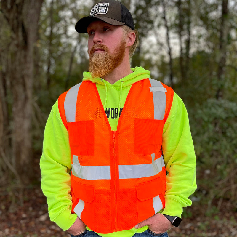 Man pictured, wearing a We Workin Safety Orange color vest, pictured from the front. Woods in the background. Also wearing a WW standard patch hat with Everybody Wants the Money...tagline.