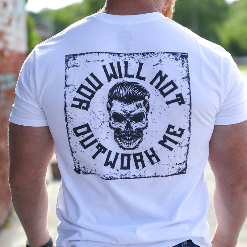 Man pictured from back wearing a We Workin graphic tee in Bright White color. Short sleeve shirt imprinted with a We Workin designed graphic—full back image with the text "You Will Not Outwork Me" encircling a skull on a distressed square pattern background (printed in black ink.)
