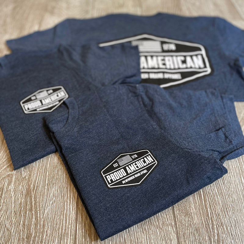 Front "pocket area" of 3 men's short sleeved t-shirts in Navy blue, folded on a tile background. "PROUD AMERICAN. EST 1776." design is printed small, pocket-sized in black and white ink.
