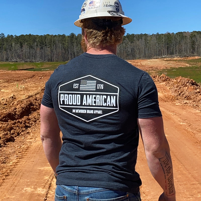 Man pictured, outdoors, from back wearing a WEWORKIN Brand Apparel Navy Blue Graphic Tee. "PROUD AMERICAN. EST 1776." design is printed in bold on full back width in black and white ink.