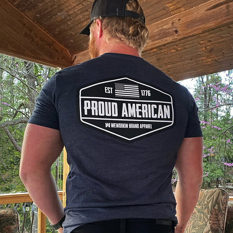 Man pictured from back wearing a WEWORKIN Brand Apparel Navy Blue Graphic Tee. "PROUD AMERICAN. EST 1776." design is printed in bold on full back width in black and white ink. Also wearing a WEWORKIN Script logo Retro Trucker hat.