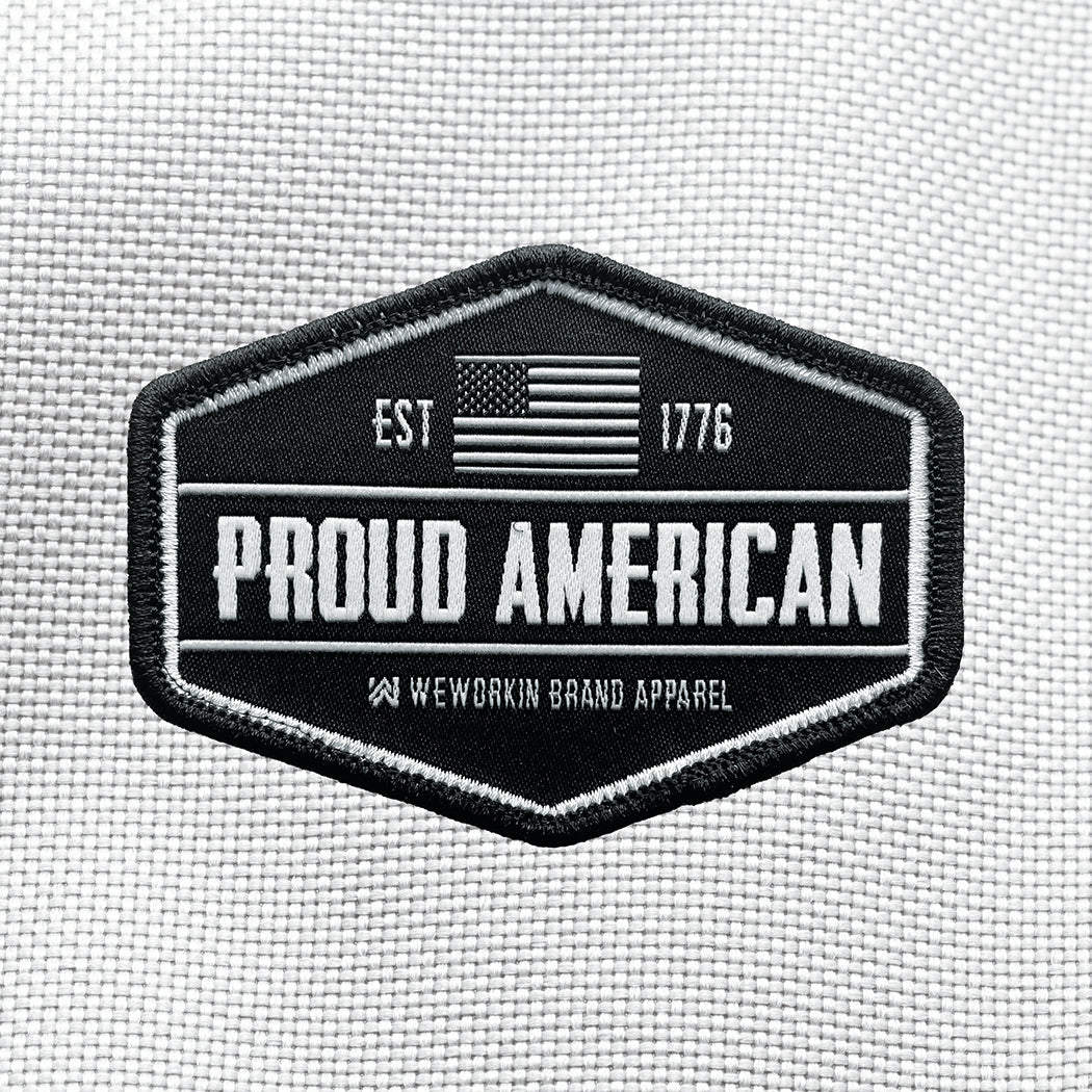 "PROUD AMERICAN" text centered large on velcro-back patch (both the hook and loop velcro sides included). [2] thread colors for the design (grey and white) on a black woven background, with black merrowed border. EST 1776 and US flag centered at top of patch.3.5" wide Woven patch displayed on a grey canvas background.
