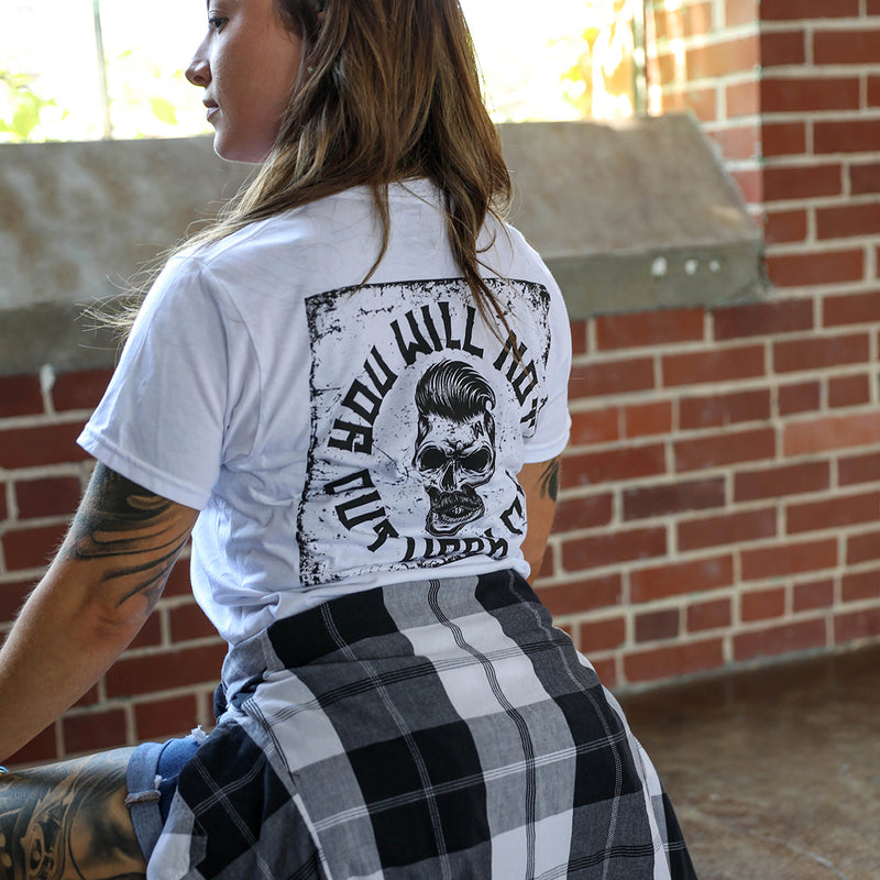 Woman pictured from back, kneeling down and wearing a We Workin graphic tee in Bright White color. The white short sleeve shirt is imprinted with a We Workin graphic—full back image with the text "You Will Not Outwork Me" encircling a skull on a distressed square pattern background (printed in black ink.) 