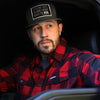 Man pictured from front wearing a We Workin long sleeve Flannel buttoned shirt in Red/Black Buffalo Plaid, sitting in the driver's seat of a vehicle. A small/rectangular black with white thread WEWORKIN BRAND woven tag is sewn on the left pocket flap. He is also wearing a We Workin Flat Bill Snapback Hat in Charcoal/Black with a large Patch that says "Everybody Want$ the Money, Nobody Wants the WORK." in grey/white thread on Black woven background with white merrowed-edge.