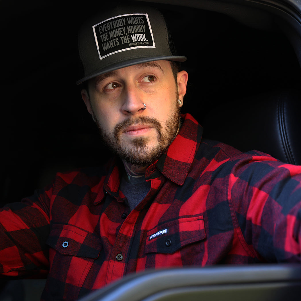 Man pictured from front wearing a We Workin long sleeve Flannel buttoned shirt in Red/Black Buffalo Plaid, sitting in the driver's seat of a vehicle. A small/rectangular black with white thread WEWORKIN BRAND woven tag is sewn on the left pocket flap. He is also wearing a We Workin Flat Bill Snapback Hat in Charcoal/Black with a large Patch that says "Everybody Want$ the Money, Nobody Wants the WORK." in grey/white thread on Black woven background with white merrowed-edge.