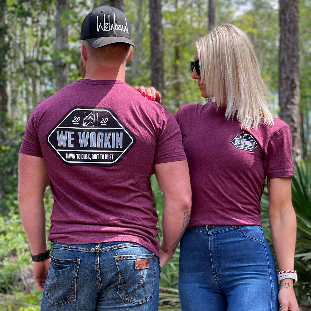 Man (back) and woman (front) wearing a We Workin graphic tee in Heather Burgundy Rust color. Short sleeve shirt printed with We Workin designed emblem graphic—"WE WORKIN. DAWN TO DUSK, DIRT TO RUST" printed boldly across the full back (in black, white and grey ink). Man is also wearing a stealth black WW Classic Retro Trucker snapback with the WE WORKIN script logo embroidered in white thread on front panels.