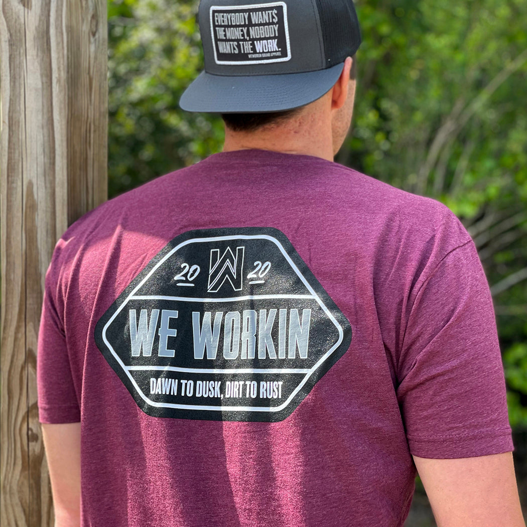 Man wearing a We Workin graphic tee in Heather Burgundy Rust color. Short sleeve shirt printed with We Workin designed emblem graphic—"WE WORKIN. DAWN TO DUSK, DIRT TO RUST" printed boldly across the full back (in black, white and grey ink). Man is also wearing a charcoal grey/black WW Flatbill Patch Snapback with text "Everybody Wants the Money, Nobody Wants the WORK" on patch.