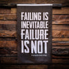 WeWorkin Brand "FAILING IS INEVITABLE—FAILURE IS NOT" flag displayed on a rough wood wall. Each flag measures approximately 3'W x 5'H. The flag has a black background with all white letters. White, double-stitched, thicker top edge for durability, (2) gold grommets (one at top left and one at top right corners).