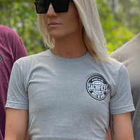 Woman pictured from back, outdoors, wearing a Men's We Workin Athletic Grey short sleeve Graphic Tee. "SACRIFICES MUST BE MADE." design is printed on the left chest "pocket area" in black and white ink.