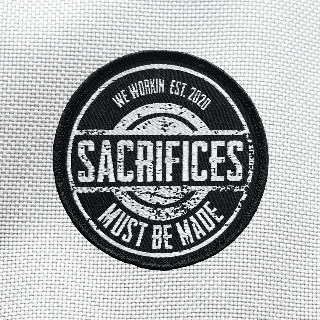 "SACRIFICES" text large and centered on patch, "MUST BE MADE" circling the bottom border, on a velcro-backed patch (both the hook and loop velcro sides included). "WE WORKIN EST. 2020" circles the top border. [1] thread color for the design (white) on a black woven background, with black merrowed border. 3.25" wide circular Woven patch displayed on a grey canvas background.