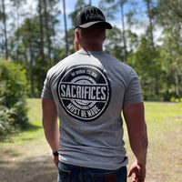 Man pictured from back, outdoors, wearing a We Workin Athletic Grey Graphic Tee. "SACRIFICES MUST BE MADE." design is printed boldly on full back width in black and white ink. He is also wearing a We Workin retro trucker hat in Stealth Black with our embroidered script logo .