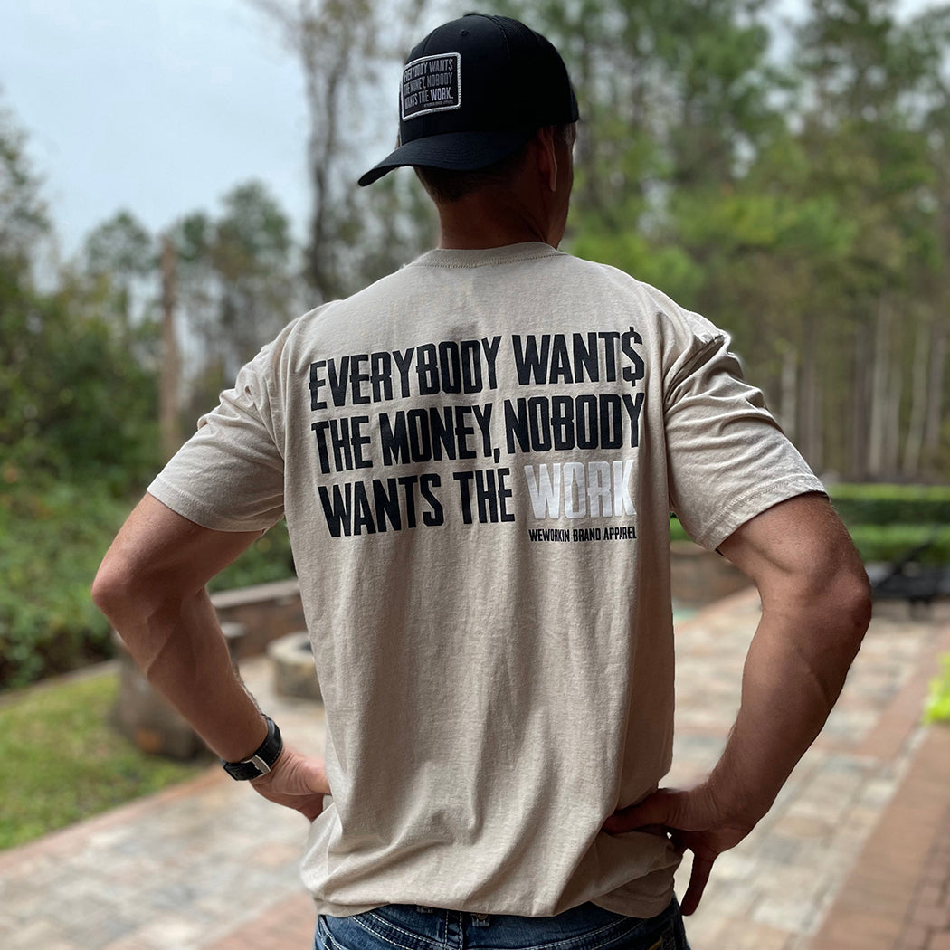 Man pictured from back wearing a WW Desert Sand color Graphic Tee. "EVERYBODY WANT$ THE MONEY, NOBODY WANTS THE WORK" printed in bold on full back width in black ink, except the word WORK is printed in white for emphasis. (WEWORKIN BRAND APPAREL printed small and lower right just below the full back imprint.) Also wearing a WW Small Patch EWTM hat backwards.