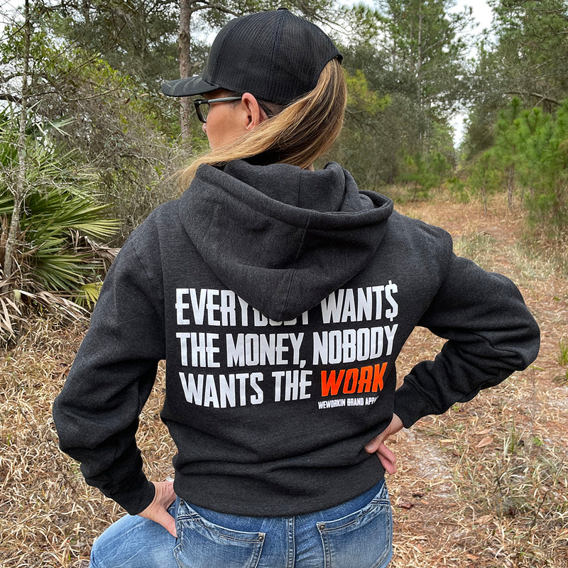 Woman pictured from back wearing a WW HARD Charcoal Grey Hoodie. "EVERYBODY WANT$ THE MONEY, NOBODY WANTS THE WORK" printed in bold on full back width in white ink, except the word WORK is printed in neon orange for emphasis. (WEWORKIN BRAND APPAREL printed small and lower right just below the full back imprint.) Banded cuffs and waist. Hood down on back.