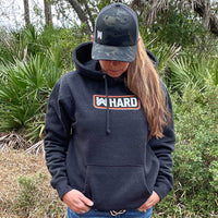 Woman pictured from front wearing a WW HARD Charcoal Grey Hoodie. WW HARD "patch-style" icon printed in neon orange and white, small on center chest. Banded cuffs and waist. (Also wearing a WW Black camo retro trucker hat)