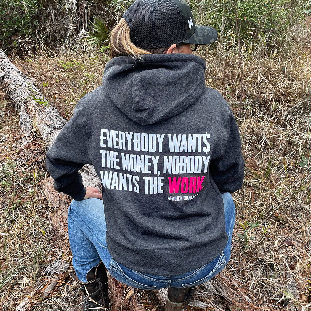 Woman pictured from back wearing a WW HARD Charcoal Grey Hoodie. "EVERYBODY WANT$ THE MONEY, NOBODY WANTS THE WORK" printed in bold on full back width in white ink, except the word WORK is printed in neon pink for emphasis. (WEWORKIN BRAND APPAREL printed small and lower right just below the full back imprint.) Banded cuffs and waist.