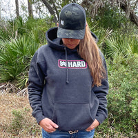 Woman pictured from front wearing a WW HARD Charcoal Grey Hoodie. WW HARD "patch-style" icon printed in neon pink and white, small on center chest. Banded cuffs and waist.  Also wearing a WeWorkin Black Camo Retro Trucker snapback.