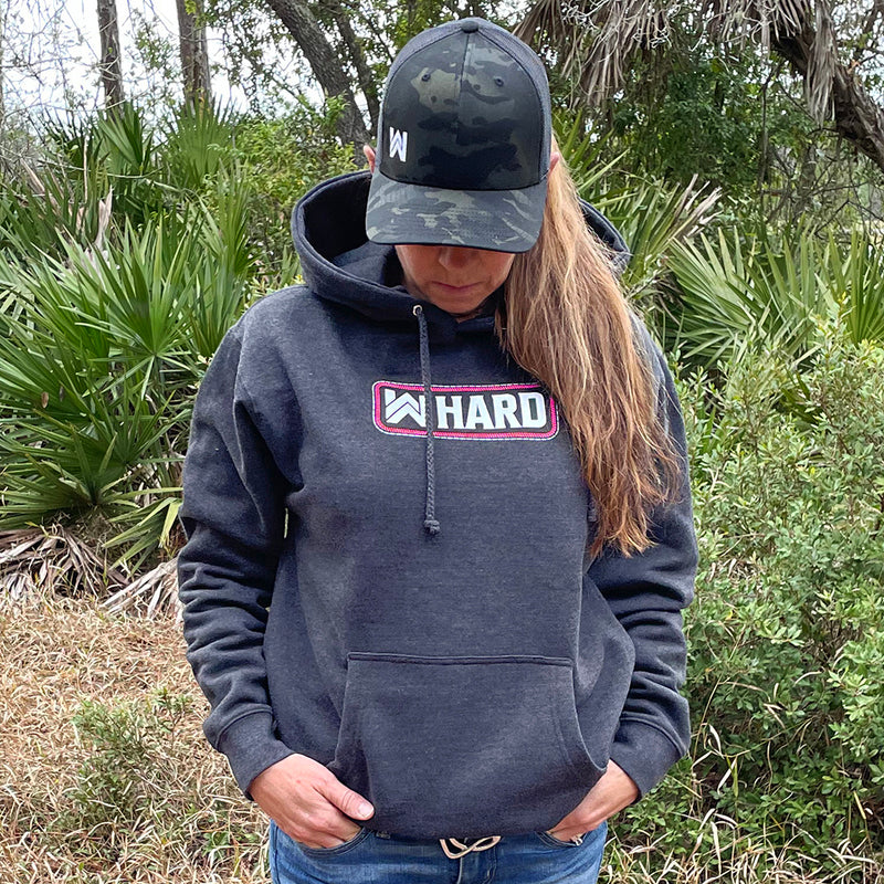 Woman pictured from front wearing a WW HARD Charcoal Grey Hoodie. WW HARD "patch-style" icon printed in neon pink and white, small on center chest. Banded cuffs and waist.  Also wearing a WeWorkin Black Camo Retro Trucker snapback.