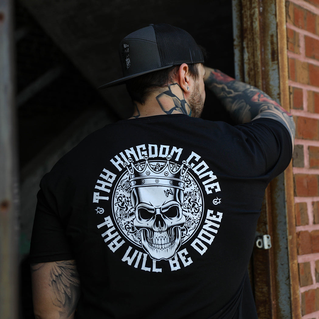 Man pictured from back wearing a We Workin graphic tee in Dark Black color. Short sleeve shirt imprinted with a WeWorkin designed graphic—the text "Thy Kingdom Come Thy Will Be Done" encircling a skull with crown on a nordic circular pattern (printed in white ink.) He is also wearing the WeWorkin Brand 7-panel Charcoal Grey/Black Skull hat. 