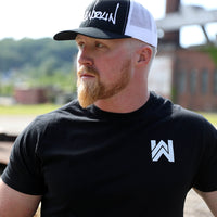 Man pictured from front, head turned to the right, wearing the WeWorkin Retro Trucker Hat in Black/White with embroidered WW script logo across the front in white thread. He is also wearing a WeWorkin black tee with the WW icon logo printed small on the left chest in white ink.