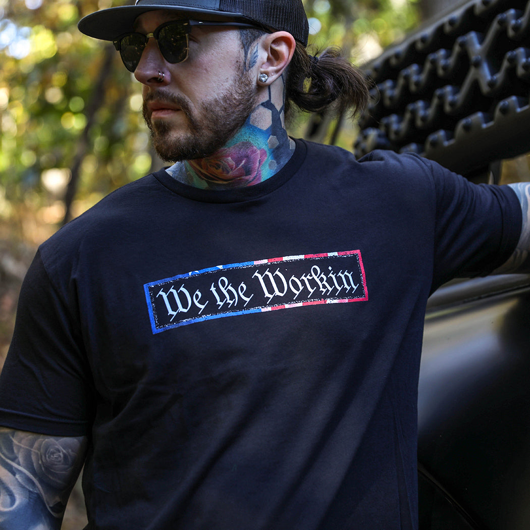 Man outdoors wearing a WE WORKIN men's short sleeved t-shirt in Black. "We the Workin" text with textured American flag patterned rectangle border printed around it, on center chest in full color. Also wearing a WW flat bill snapback in grey/black with LG patch.