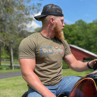 Man outdoors wearing a WE WORKIN men's short sleeved t-shirt in Desert Green. "We the Workin" text with textured American flag patterned rectangle border printed around it, on center chest in full color. Also wearing a WW Trucker Snapback Patch Hat in Black.