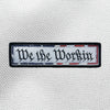 "We The Workin" text and red/white/blue flag border design centered on a velcro-backed patch (both the hook and loop sides provided). [4] thread colors for the design (red, white, blue and black) on a white woven background, with black merrowed border. 4.5" wide Woven patch displayed on a grey canvas background.