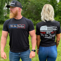 Man and woman pictured outdoors, both wearing the WW "We the Workin—Built by We the People" tee in Black. He is standing forward and she is showing the back of the tee.