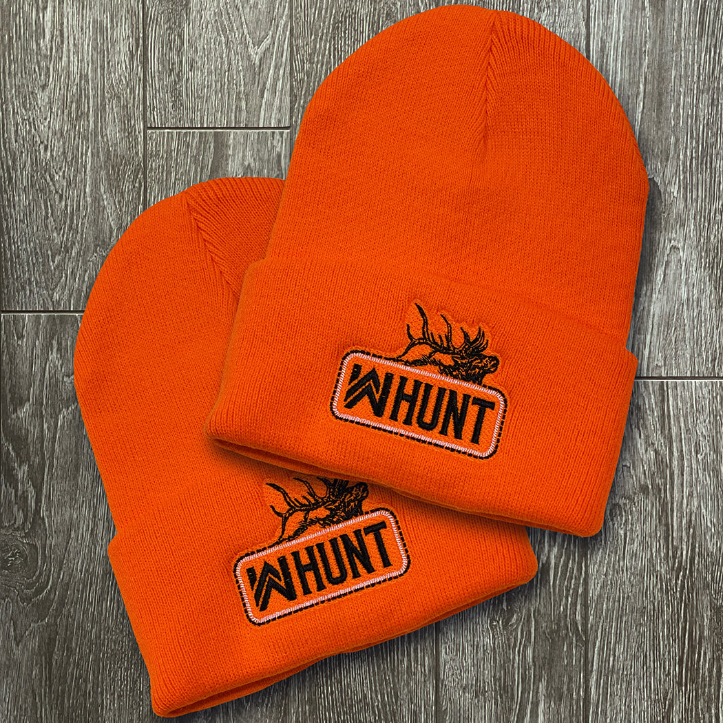 Two WW HUNT Neon Safety Orange cuffed beanies on a grey tile background. Embroidered with WW HUNT and ELK image in our signature "patch" style on the folded cuff (WW icon/text/elk graphic in black thread, outer outline in black thread, inner "rope" in white thread). 
