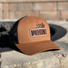 Picture of our WW HUNT trucker hat in duotone Caramel and black, placed on a stone fire pit. Embroidered with WW HUNT and ELK in our signature "patch" style across the front (WW icon/text/elk graphic in black thread, outer outline in black thread, inner "rope" in white thread). Front 2 panels and bill are caramel color, side/back mesh panels are black.