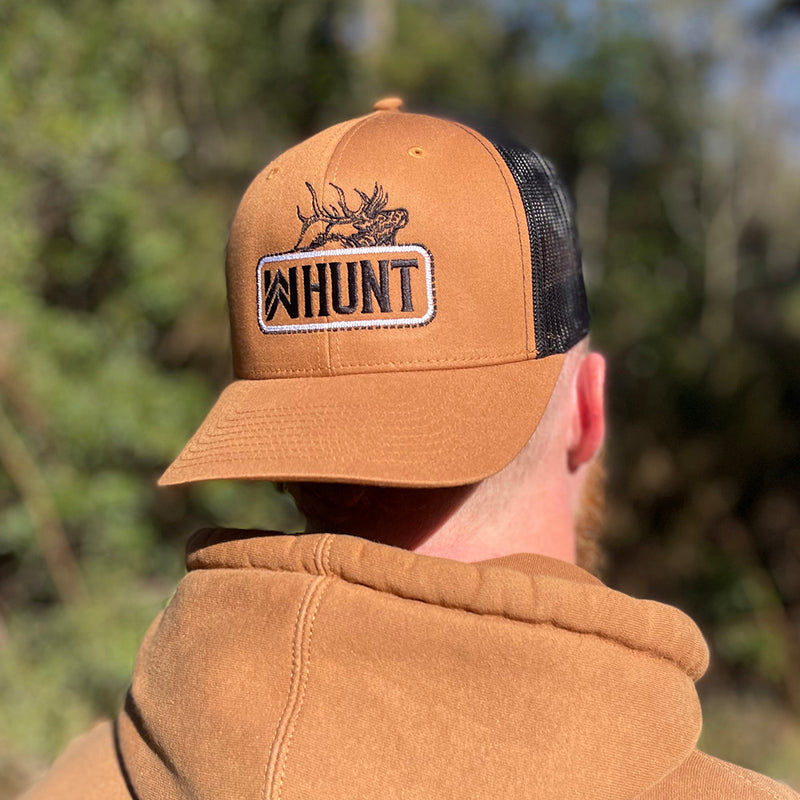 Man wearing a WW HUNT trucker hat in duotone Caramel and black, backwards. Embroidered with WW HUNT and ELK in our signature "patch" style across the front (WW icon/text/elk graphic in black thread, outer outline in black thread, inner "rope" in white thread). Front 2 panels and bill are caramel color, side/back mesh panels are black color.