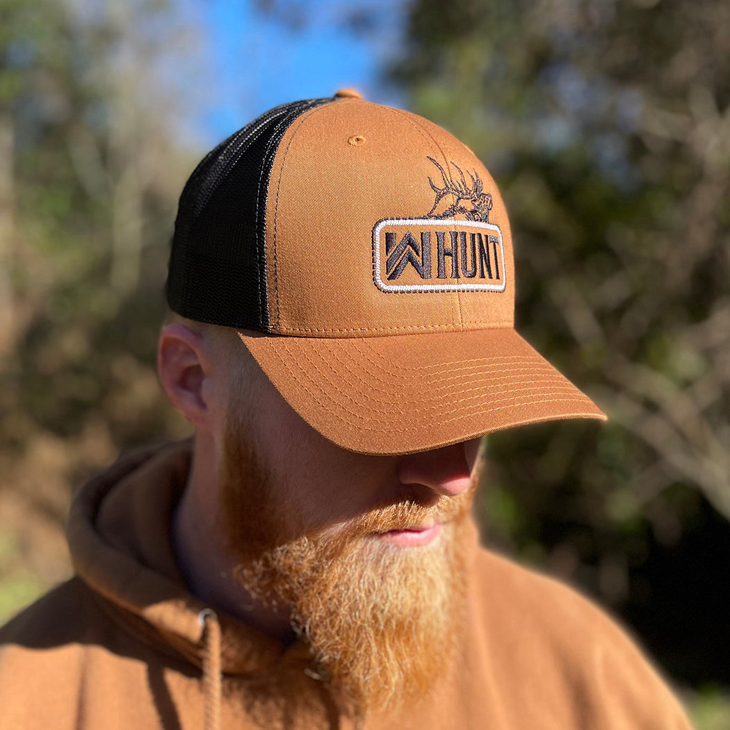 Man wearing a WW HUNT trucker hat in duotone Caramel and black. Embroidered with WW HUNT and ELK in our signature "patch" style across the front (WW icon/text/elk graphic in black thread, outer outline in black thread, inner "rope" in white thread). Front 2 panels and bill are caramel color, side/back mesh panels are black color.