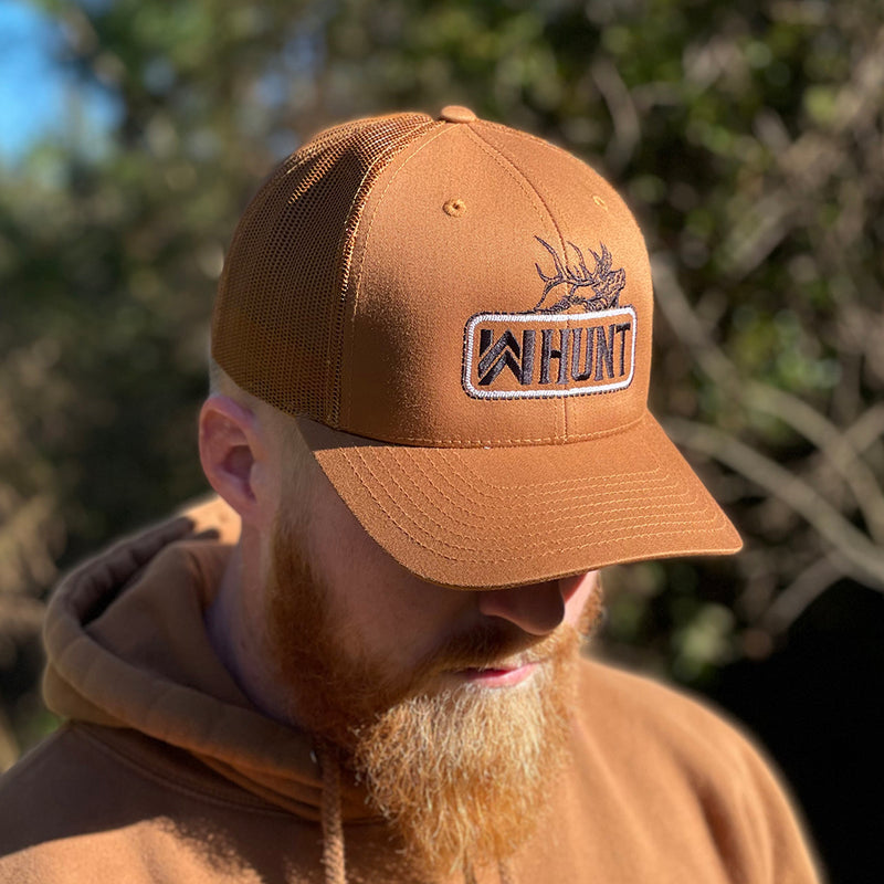 Man wearing a WW HUNT trucker hat in Caramel color, outdoors. Embroidered with WW HUNT and ELK in our signature "patch" style across the front (WW icon/text/elk graphic in black thread, outer outline in black thread, inner "rope" in white thread). Front 2 panels and bill are caramel color, side/back mesh panels are caramel color.