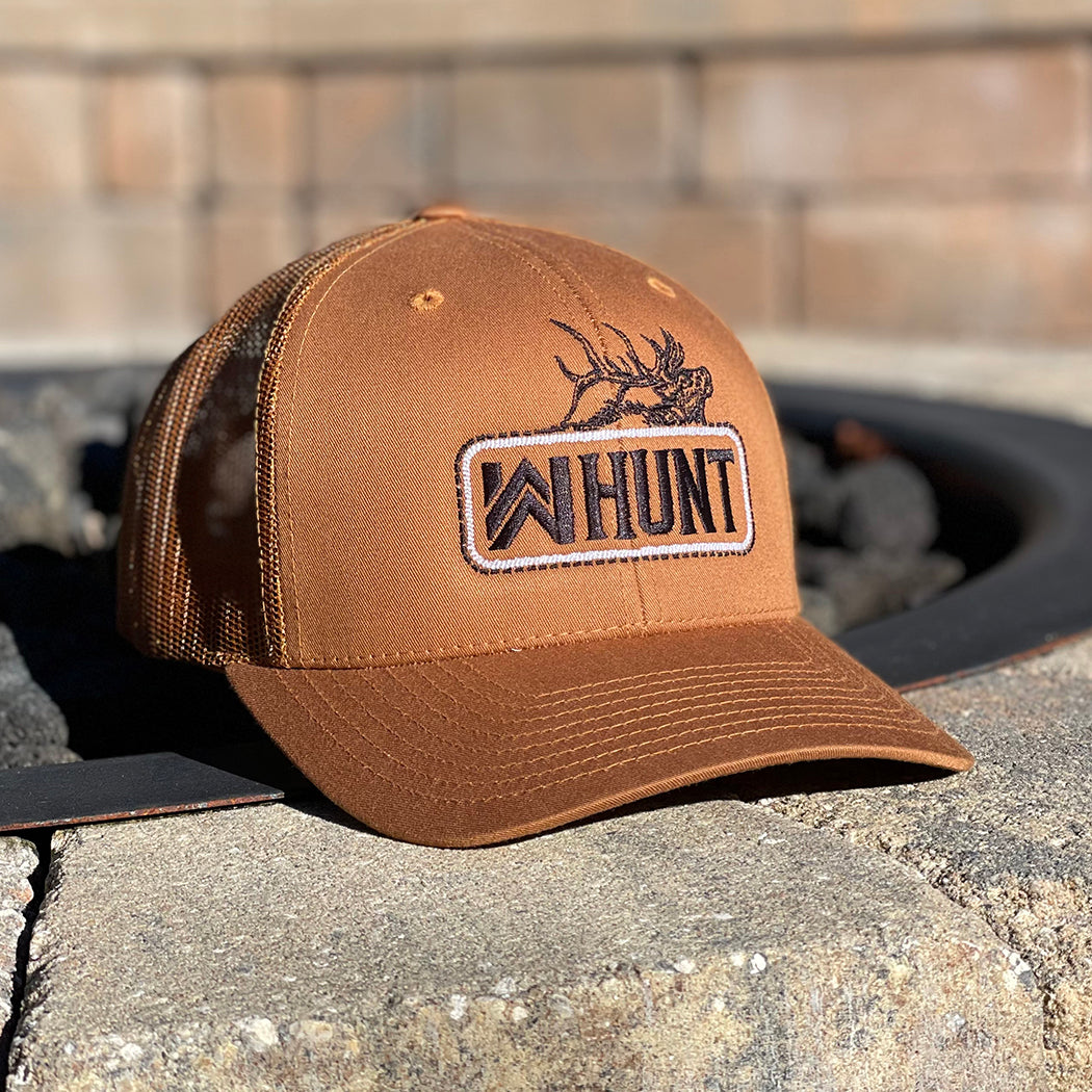 Picture of our WW HUNT trucker hat in Caramel color, placed on a stone fire pit. Embroidered with WW HUNT and ELK in our signature "patch" style across the front (WW icon/text/elk graphic in black thread, outer outline in black thread, inner "rope" in white thread). Front 2 panels and bill are caramel color, side/back mesh panels are caramel color.