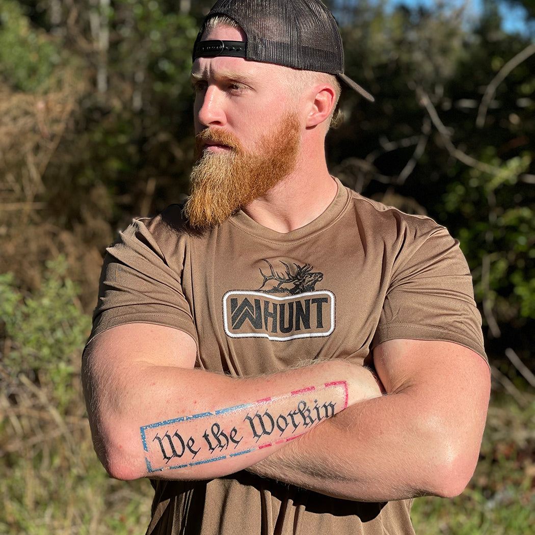 Man pictured outdoors, from the front, in a WE WORKIN ultralight weight, woodland brown short-sleeved tee—center chest screenprinted with the WW HUNT and ELK image in our signature "patch" style (WW icon/text/elk graphic in black ink, outer outline in black ink, inner "rope" in white ink).