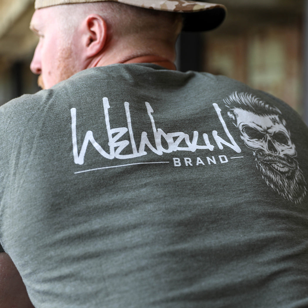 Man pictured from back wearing a We Workin graphic tee in Heather Military Green color. Short sleeve shirt imprinted with a WeWorkin designed graphic—the WeWorkin Brand Skull icon, large across the back (printed in white ink.) He is also wearing the WeWorkin Arid Tan Camo Retro Trucker hat. 