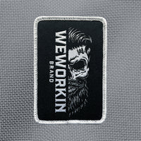 "WEWORKIN BRAND" text running vertically along a bearded half skull on a velcro-backed patch (both the hook and loop sides provided). [1] thread color for the design (white) on a black woven background, with white merrowed border. 3.5" tall Woven patch displayed on a grey canvas background.