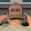 Man wearing a Coyote Brown and Black Retro Trucker hat backwards. Embroidered with WW HARD "patch" style across the front (WW HARD icon/text in black thread, outer outline in orange thread, inner outline in black thread). Front 2 panels and bill are coyote brown, side/back mesh panels are black.