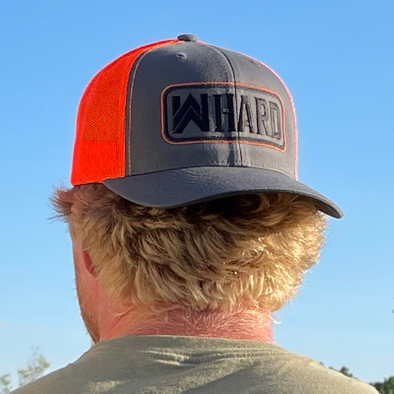 Man wearing a Neon Orange and Grey Retro Trucker hat backwards. Embroidered with WW HARD "patch" style across the front (WW HARD icon/text in black thread, outer outline in orange thread, inner outline in black thread). 